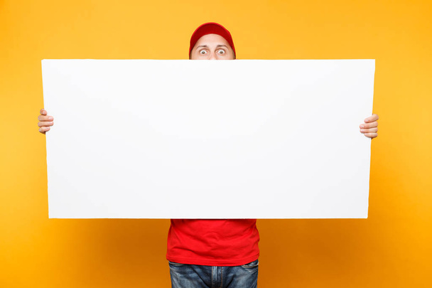Delivery man in red uniform isolated on yellow orange background. Male employee courier in cap, t-shirt holding big white empty blank billboard with copy space, place for text image. Service concept - Fotografie, Obrázek