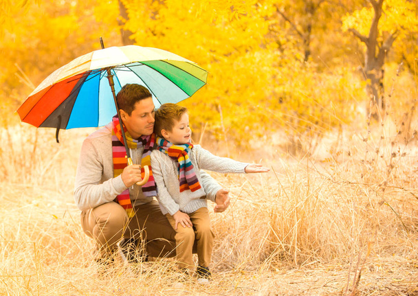 A little boy and his father are under a colorful umbrella. Family in warm clothes and colorful scarves. the kid catches the rain drops with his palm - Foto, afbeelding