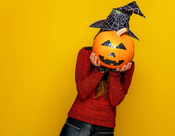 young woman in halloween witch costume holding sad jack-o-lantern pumpkin in front of face on yellow background - Фото, изображение