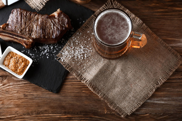Mug of delicious beer with grilled steak on wooden table - Fotoğraf, Görsel