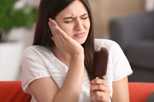 Νεαρή γυναίκα με ευαίσθητα δόντια και κρύο παγωτό στον καναπέ στο σπίτι - Φωτογραφία, εικόνα