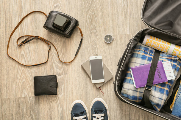Открытый чемодан с одеждой и дорожными предметами на деревянном фоне
 - Фото, изображение