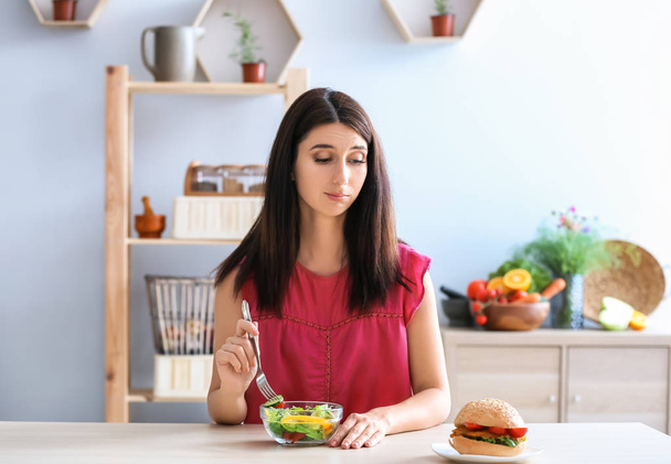 Женщина с вкусным бургером и свежим салатом в помещении. Выбор между здоровой и нездоровой пищей
 - Фото, изображение