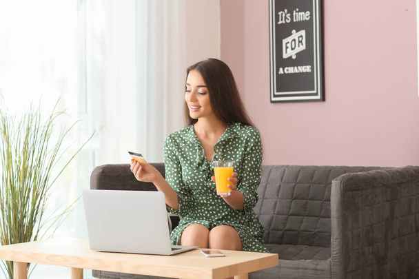 Nuori nainen, jolla on luottokortti ja lasi mehua, istuu sohvalla kotona. Verkkokauppa
 - Valokuva, kuva