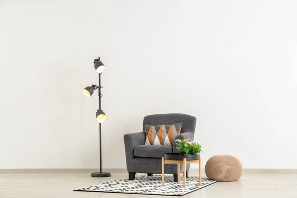 Comfortabele fauteuil met poef, tabel en vloerlamp in de buurt van lichte muur - Foto, afbeelding