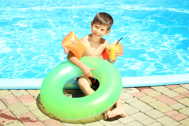 Lindo niño pequeño con anillo inflable y vaso de jugo descansando cerca de la piscina
 - Foto, Imagen