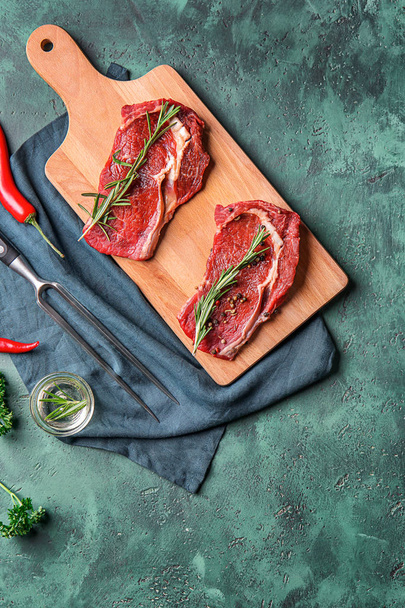 Wooden board with raw meat, rosemary and spices on color textured background - 写真・画像