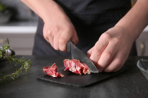 Man cutting raw meat on slate plate in kitchen - Fotoğraf, Görsel