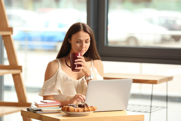 Beautiful young woman with cup of sweet cocoa drink using laptop in cafe - Photo, Image