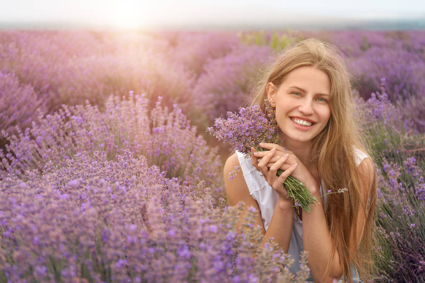 Beautiful young woman with bouquet of lavender in field on summer day - Фото, изображение