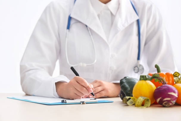 Nutricionista femenina sentada a la mesa con portapapeles y productos saludables sobre fondo blanco
 - Foto, Imagen