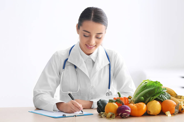 Nutricionista femenina sentada a la mesa con portapapeles y productos saludables sobre fondo blanco
 - Foto, imagen