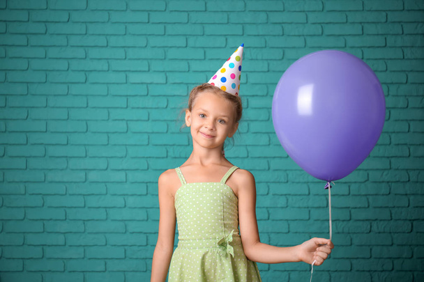 Linda niña en sombrero de cumpleaños y con globo contra la pared de ladrillo de color
 - Foto, Imagen