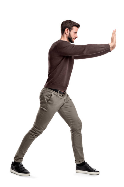 A bearded man in casual clothes tries to push a heavy object with both arms with one leg put in front for balance. - Photo, image