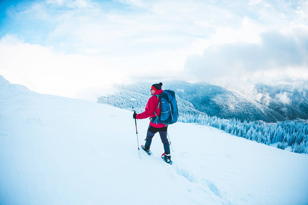 Ein Mann in Schneeschuhen und Trekkingstöcken in den Bergen. Winterreise. Klettern eines Bergsteigers gegen einen schönen Himmel mit Wolken. Aktiver Lebensstil. den Berg hinauf durch den Schnee. - Foto, Bild