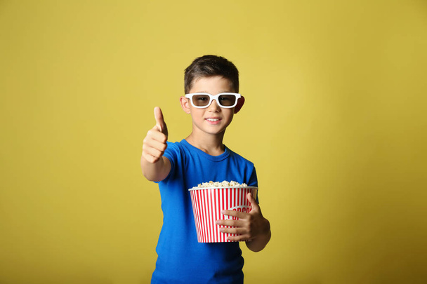 Cute little boy with cup of popcorn watching movie and showing thumb-up gesture on color background - Foto, Imagem
