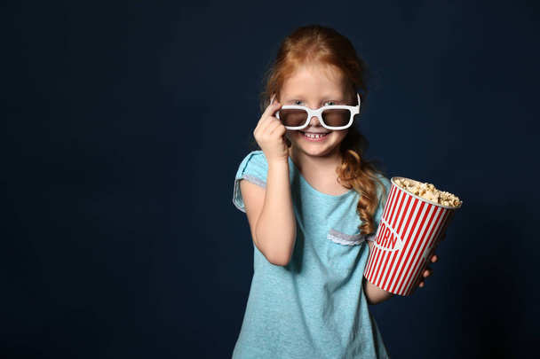 Cute little girl with cup of popcorn wearing 3D cinema glasses on dark background - Foto, immagini