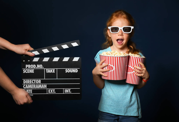 Female hands with clapper board and cute girl holding cups of popcorn on dark background - Foto, Imagen