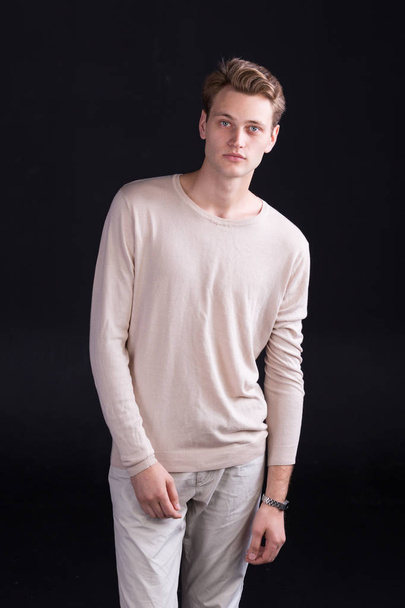 Handsome male model with blue eyes and light curlie hair in beige sweater and white pants on a dark background - Фото, зображення