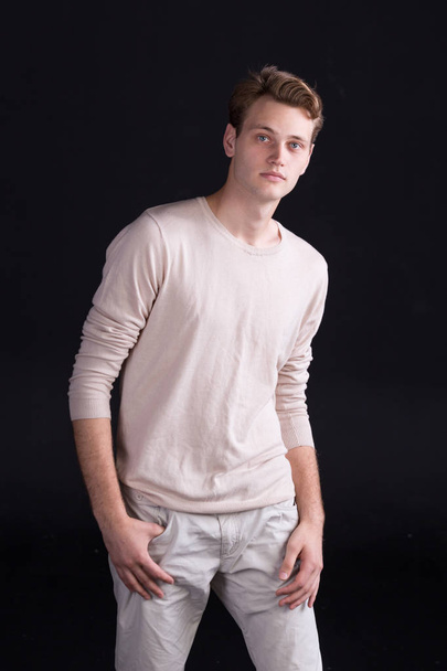 Handsome male model with blue eyes and light curlie hair in beige sweater and white pants on a dark background - Foto, immagini