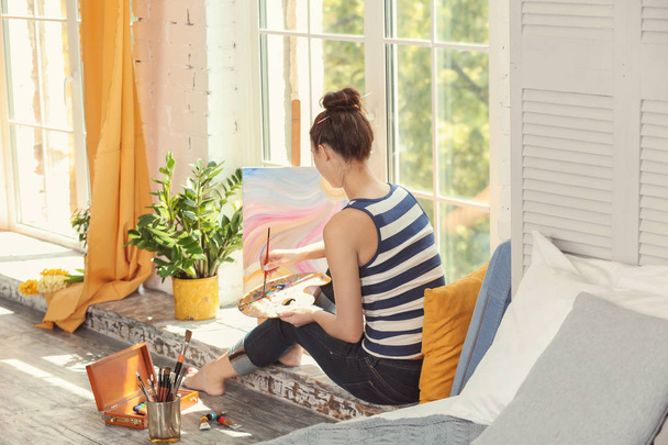 Künstlerin malt Bild während sie auf Fensterbank in Werkstatt sitzt - Foto, Bild