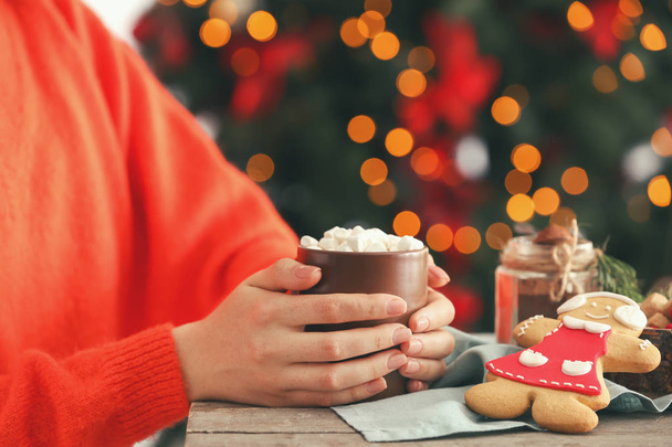 Жінка з чашкою смачного какао за столом проти розмитих різдвяних вогнів, крупним планом
 - Фото, зображення
