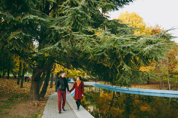 Jeune couple heureux en amour femme et homme en vêtements chauds se tenant la main en se regardant marcher sur la route près de l'eau dans le parc de la ville d'automne à l'extérieur. Amour relation famille gens style de vie concept
 - Photo, image