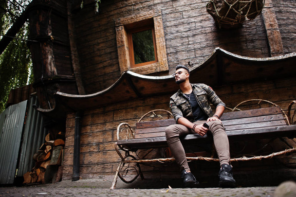 Awesome beautiful tall ararbian beard macho man in glasses and military jacket posed outdoor, sitting on bench against wooden house. - Zdjęcie, obraz