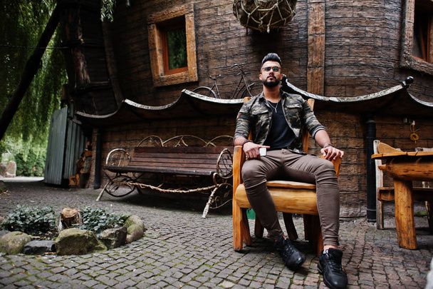 Awesome beautiful tall ararbian beard macho man in glasses and military jacket posed outdoor, sitting on chair against wooden house. - Zdjęcie, obraz