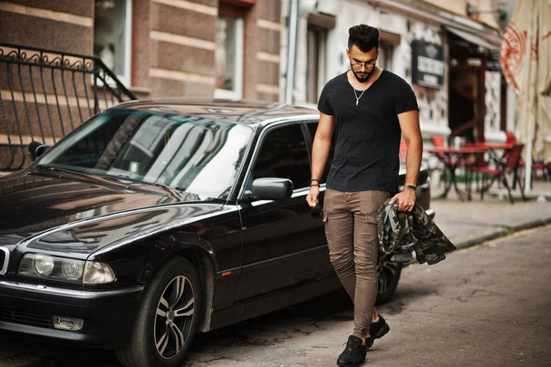 Awesome beautiful tall ararbian beard macho man in glasses and black t-shirt walking against business car. - Foto, imagen