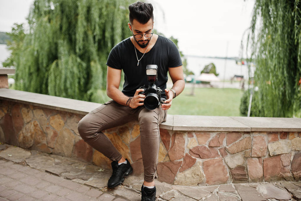 Awesome beautiful tall ararbian beard macho man photographer in glasses and black t-shirt with professional camera at hands. - Foto, Imagen