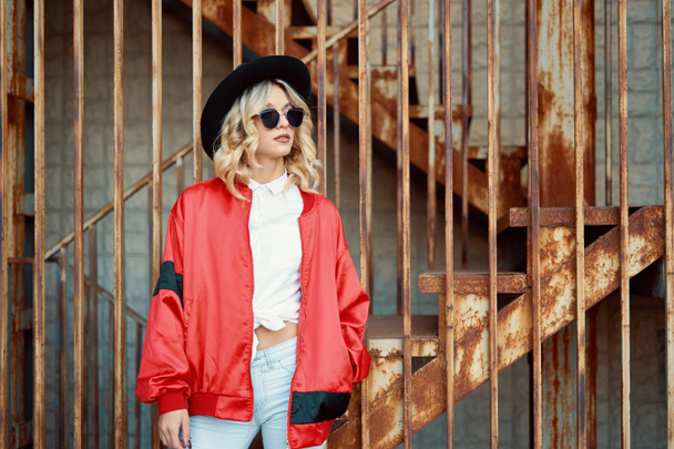 Güzel ve şık bir genç kadın paslı metal merdivenlerde, şık giysiler, kırmızı ceket, beyaz gömlek, siyah şapka ve güneş gözlüğü takıyor teşkil etmektedir. Pop kültürü. Moda hipster portre. - Fotoğraf, Görsel