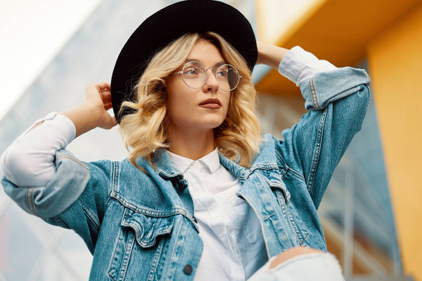 Close-up portrait of a cheerful white woman in glasses touching her hat on urban background. Photo of fashionable girl with beautiful blonde hair posing for the camera. - Photo, Image
