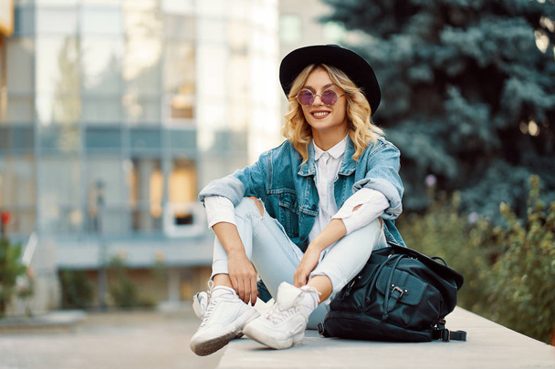 Modern lifestyle woman sitting outdoors with glass building in background. Cool young modern Caucasian female model wearing fashionable jeans, clothes, round glasses, hat and backpack. - Photo, Image