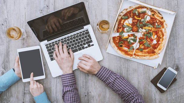 Pracownicy biurowi mają przerwę obiadową w biurze, jedzą pizzę, korzystają z laptopa i tabletu cyfrowego, leżą płasko - Zdjęcie, obraz