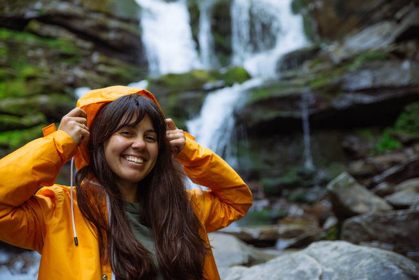 uśmiechnięta kobieta w żółty płaszcz z wodospadem na tle. pojęcie turystyki pieszej - Zdjęcie, obraz
