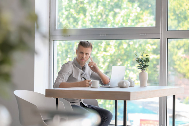 Hezký nezávislý mluví po telefonu při práci na notebooku v kavárně - Fotografie, Obrázek