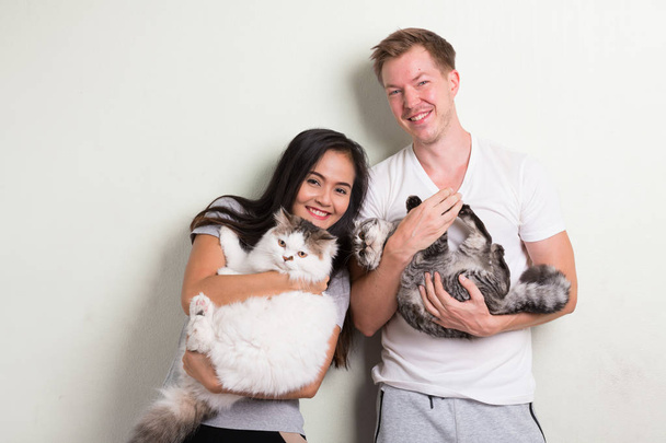 Young happy multi-ethnic couple holding two cats together against white background - Фото, зображення
