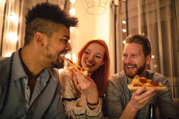 Portrait of friends enjoying eating pizza together and drinking beer at home. - Фото, изображение