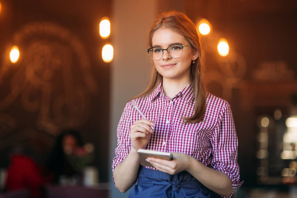 Портрет красивой дружелюбной официантки в очках с блокнотами и ручкой, готовой принять Ваш заказ
 - Фото, изображение