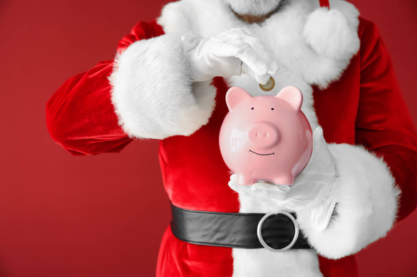 Santa Claus putting coin into piggy bank on color background, closeup - Foto, afbeelding