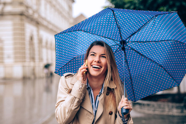A beautiful young woman talking on the phone while standing in the rain under her blue umbrella. - Zdjęcie, obraz