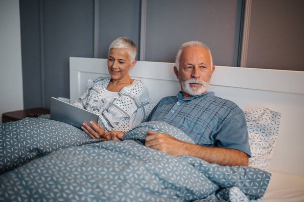 A senior happy couple lying in bed. The husband is looking out the window while the wife is happily browsing the internet on her tablet. - 写真・画像