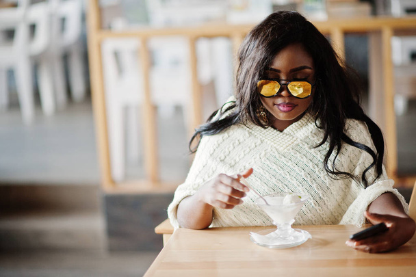 Stylish african american girl in sunglasses posed at modern cafe and eating ice cream. - Foto, Imagem