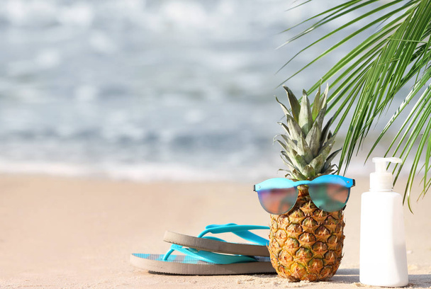Pineapple with sunglasses, flip-flops and sunscreen on sand near sea - Foto, immagini