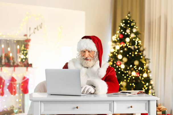 Weihnachtsmann mit Laptop sitzt am Tisch in weihnachtlich dekoriertem Zimmer - Foto, Bild