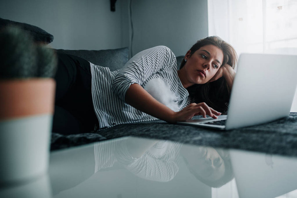 A young woman is using her laptop while lying on her couch. - Foto, Imagen