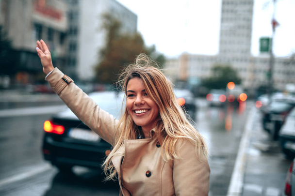A young smiling woman hailing a cab driver in a rainy city. - Fotografie, Obrázek