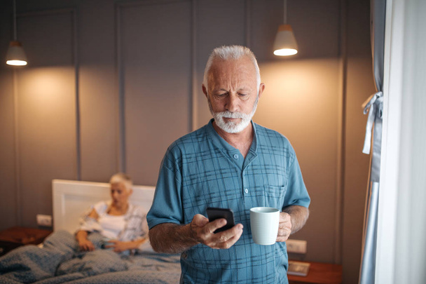 Senior man checking his phone while enjoying morning coffee. - Фото, изображение