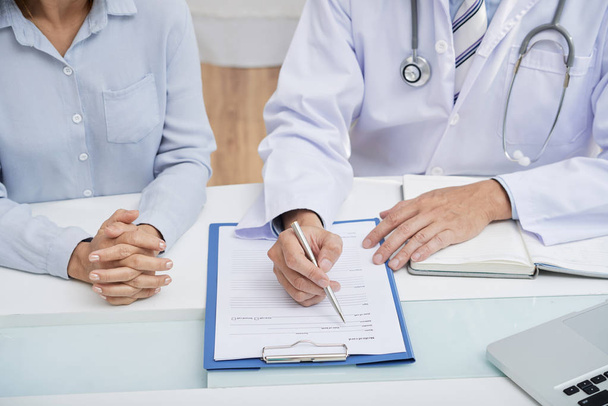 männlicher Arzt füllt Krankengeschichte aus, während er am Schreibtisch neben Patientin sitzt - Foto, Bild